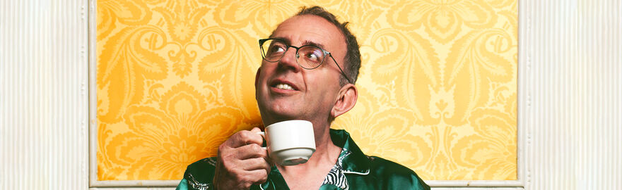 man with a cup of tea in his hands looks up in a diagonal direction whilst wearing green clothes set in a yellow and white background with patterns