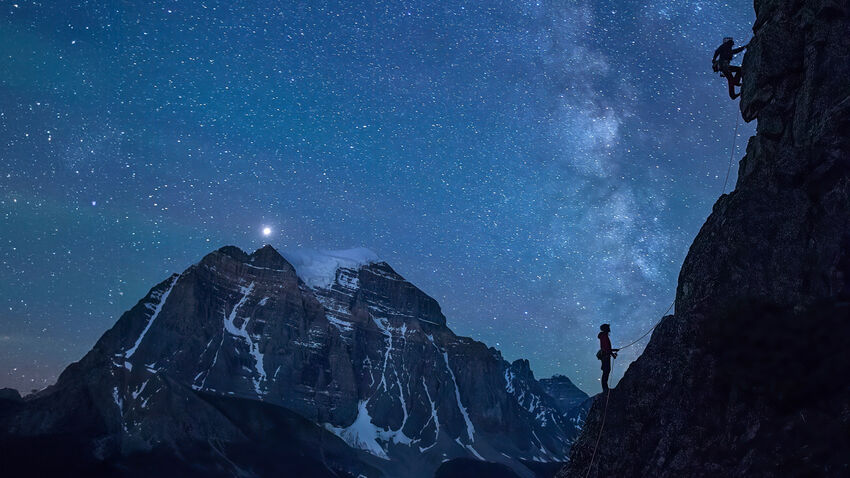 Two people climbing up a mountain at night with lots of stars and the moon glowing