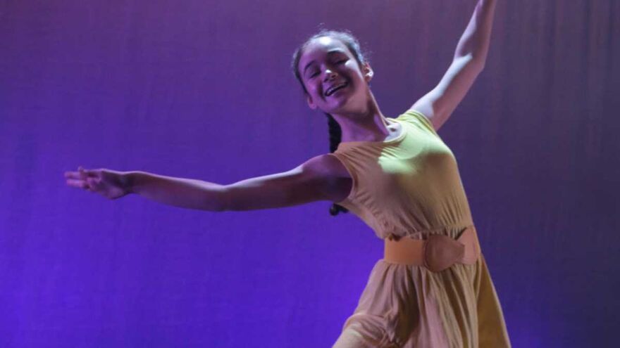 A young woman smiling on stage in a dance pose. Her arms are raised gracefully out to each side and she's wearing a simple yellow dress