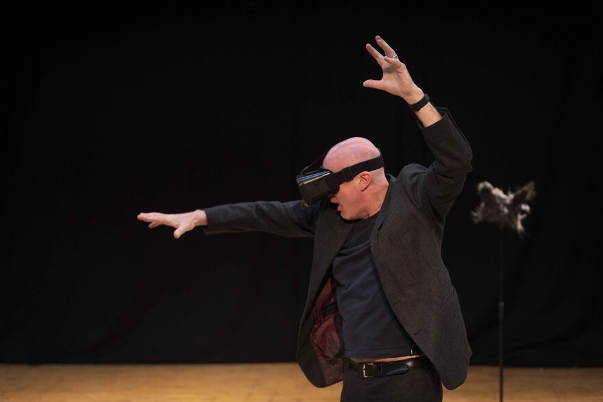 A bald white man wears a VR headset on stage, his arms are raised in dramatic fashion