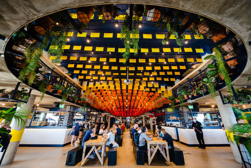 Interior of Shelter Hall, wooden tables run down the middle with black bench seating either side. People are sitting on these enjoying drinks and food. The is a big, round ceiling with orange and red bunting, and plants hanging from it.