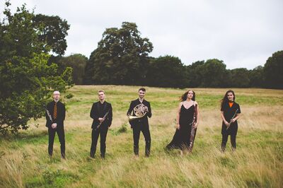 Five people in formal black attire stand in a grassy area with their instruments