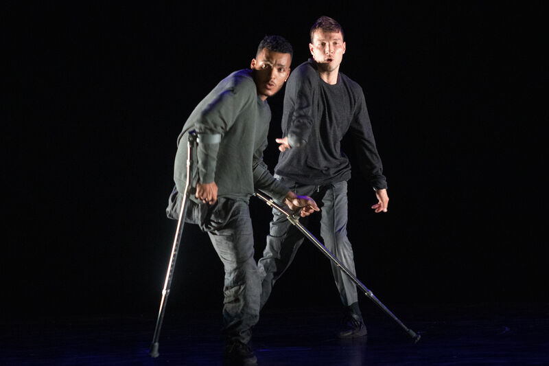 Two dancers wearing dark clothes dance on a stage. The one closest to the front is using crutches.