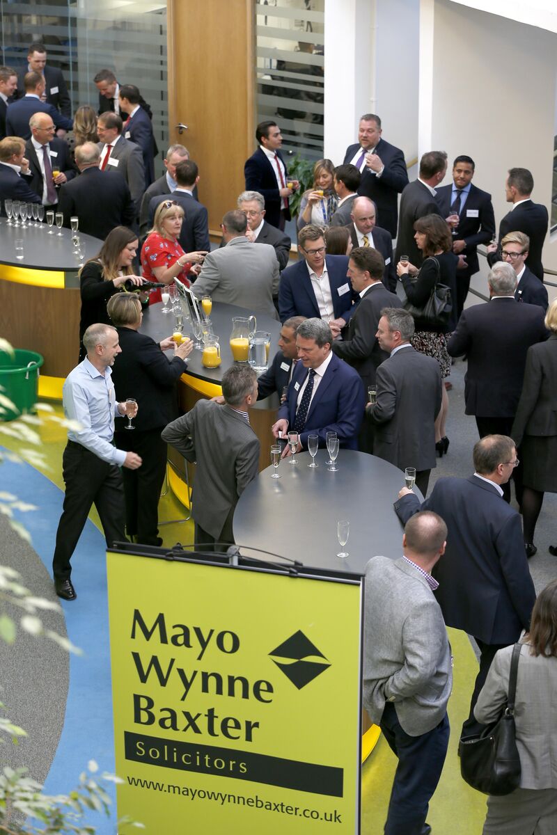 Lots of people in suits are stood around three oval tables. It is a business networking event.