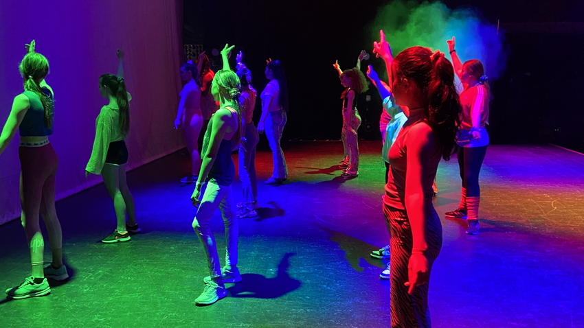 A group of children on stage with multicoloured lights pointing their hands to the sky 