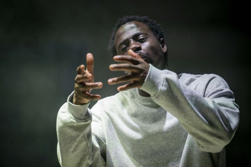 An actor wearing a grey sweater withling his hand in front of his face as though holding a spherical object