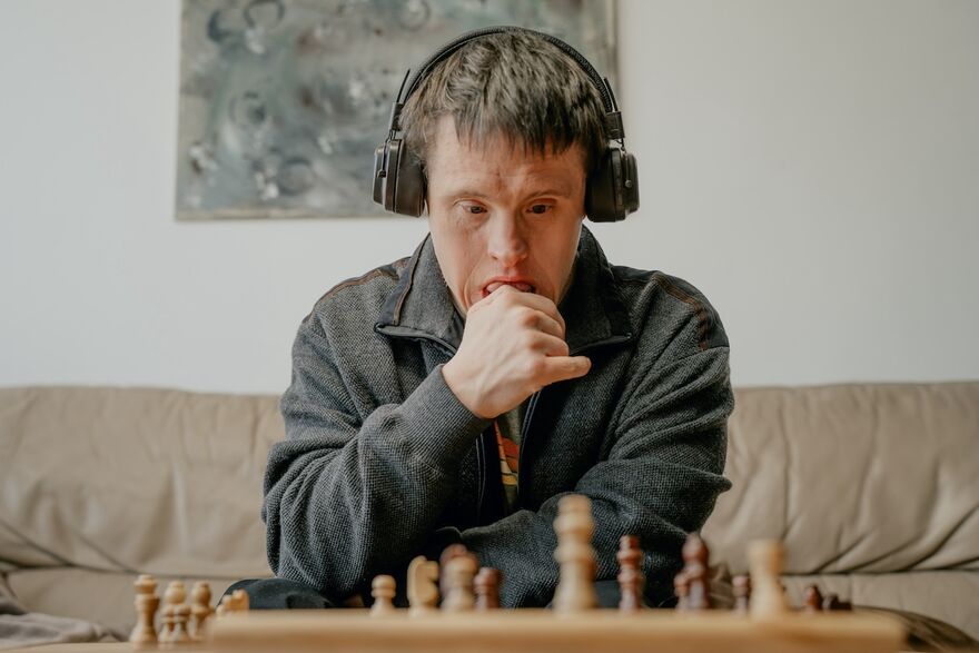 A young man with down's syndrome looks thoughtfully a game of chess in front of him. He wears a grey jumper and headphones