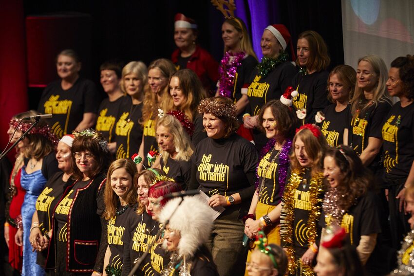 A group of singers wearing festive accessories and 'choir with no name' t-shirts