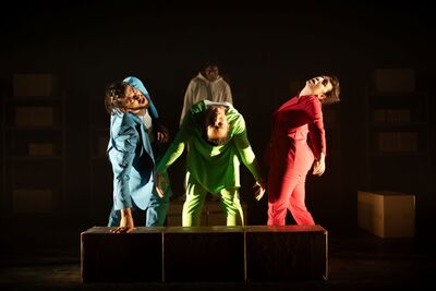 Three people in colourful suits standing in a circle leaning back. They are on a dimly lit stage with spotlights on them