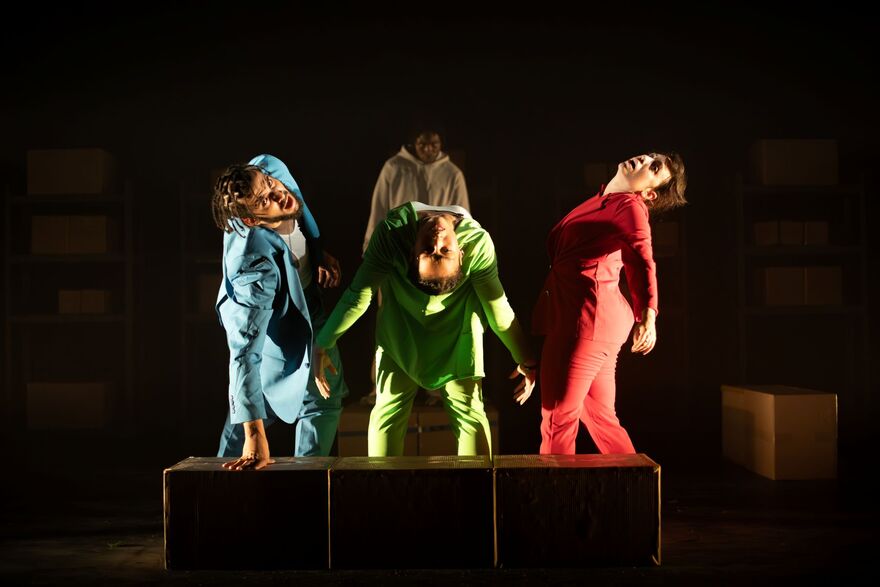 Three people in colourful suits standing in a circle leaning back. They are on a dimly lit stage with spotlights on them