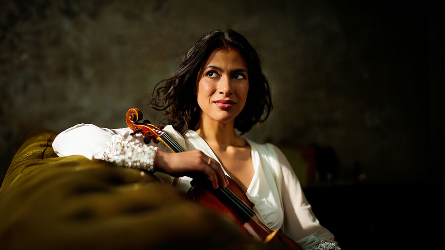 Portrait of Elena Urioste sitting on a green sofa facing the camera side-on and holding her violin.