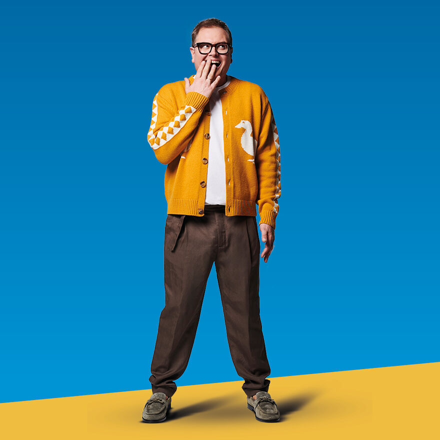 Alan Carr wearing a yellow jacket and brown trousers. He is holding his hand over his mouth and looking to the side