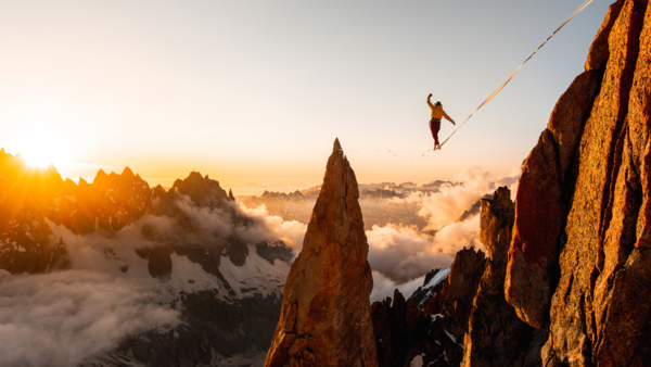 A person walks on a slackline high in the air surrounded by mountains and a misty sunrise