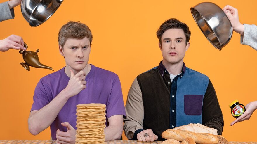 two men look directly forwards as being served food. The first man is being served pancakes, the second man is being served bread. The background is orange