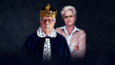 A man wearing a regal gown and crown, next to a woman wearing a pink shirt with her arms crossed 