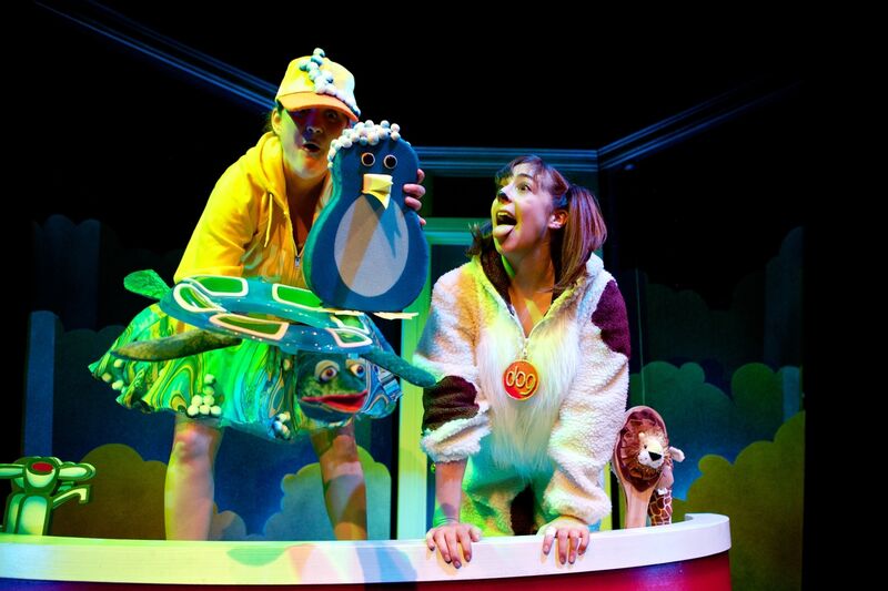 A woman dressed as a dog looks towards a penguin and turtle prop being held by another actor. Both are stood in a bath tub