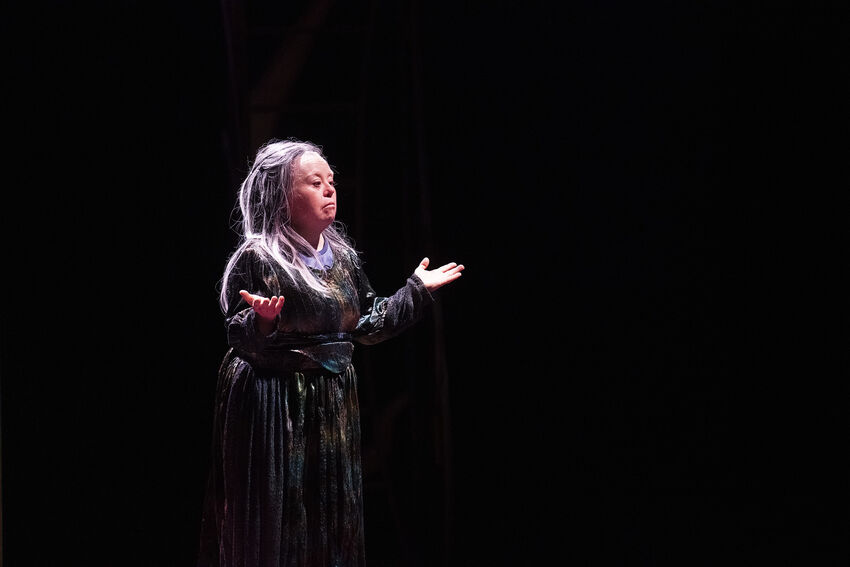 A woman with grey hair stands with her arms held out, palms open