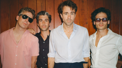 Four white men in casual shirts in front a wood pannelled wall. Two are wearing sunglasses