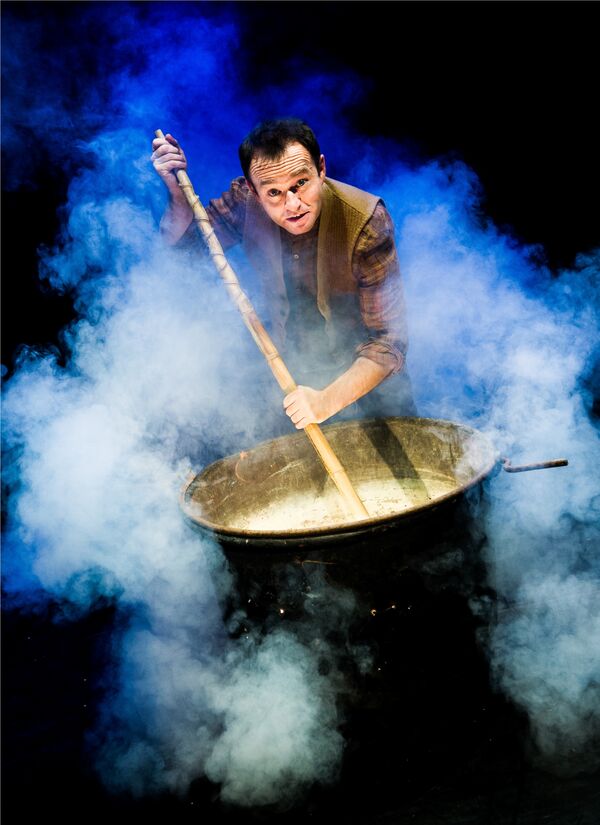 A white man in a waistcoat and shirt stands above a smoking cauldron. He is mixing its contents with a big stick.