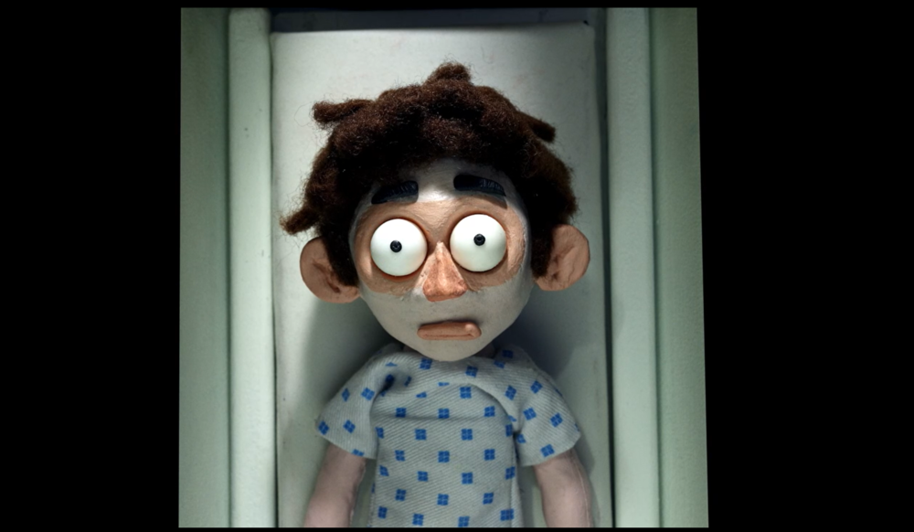 A claymation man with big eyes wearing a hopsital gown and lying in an MRI scanner