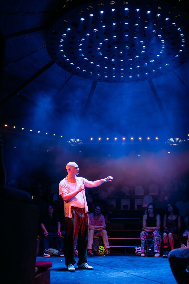 The performer stands in the middle of an in-the-round stage, with haze and moody pink and blue lighting. He is white and bald, and is speaking with his hands reached out in front of him 