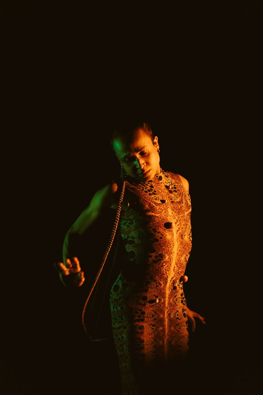 A person with a long plait wearing a long dress with a snake skin pattern. The person is looking down with their arms to their side and has a sad expression on their face
