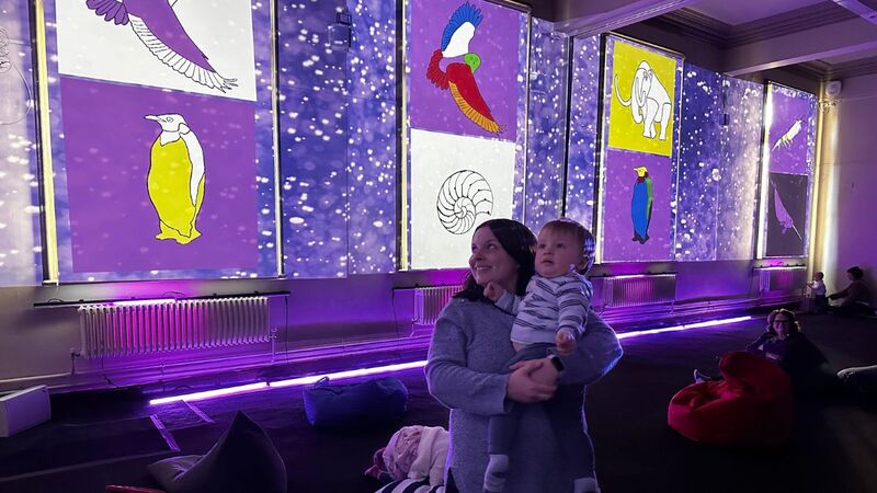 A mother holds a baby as they both look around. The room is lit by projections which cover the walls, and strip lights along the floor.