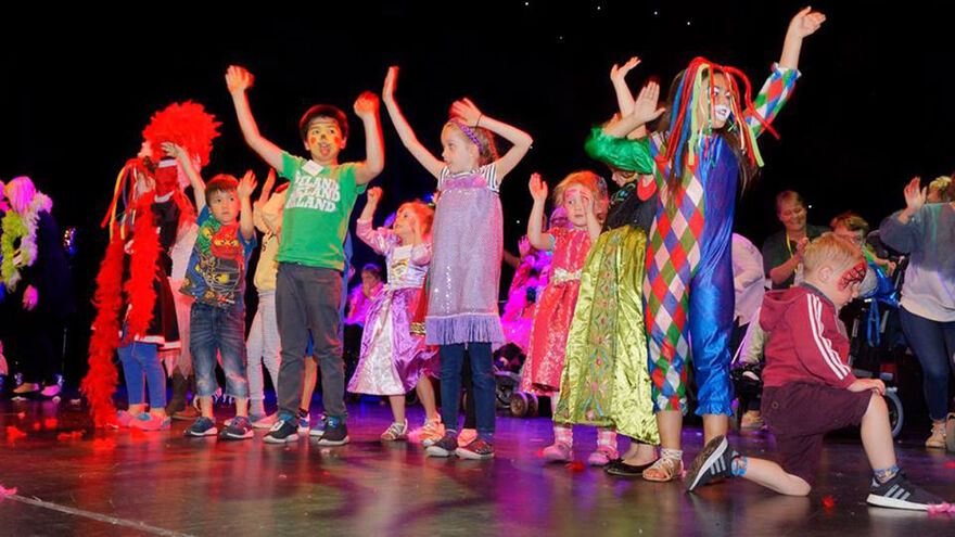 A group of children stand on stage with their arms raised up. They are all dressed in brightly coloured costumes and some have facepaint on.
