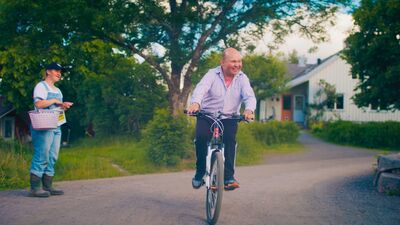 A person rides through a rural town on a bicycle. A person watches them dressed in dungarees and holding a basket