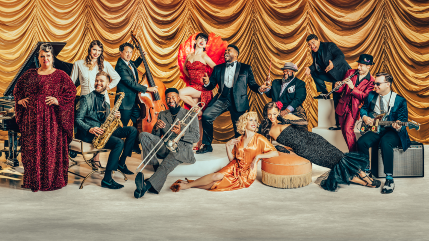A large group of musicians in glamorous ballgowns and suits, smiling and holding their instruments in front of a rouched gold silk curtain
