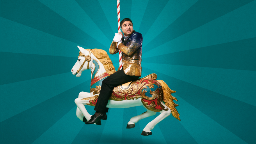 A man looking concern wearing a blue and gold sparkly blazer sitting on a carousel horse.