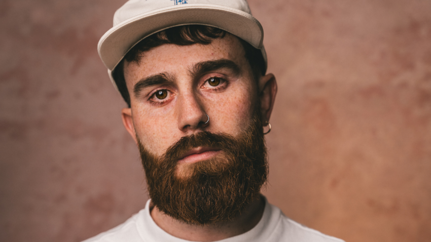 Sam Tompkins is a young white male with a beard and nose ring. He's wearing a casual white t-shirt and white baseball cap. He's looking somberly at the camera