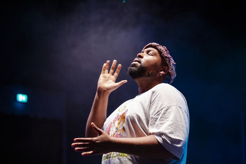 A black man stands in a spotlight, his head tilted upwards, and his hands wide apart, as if he were about to clap