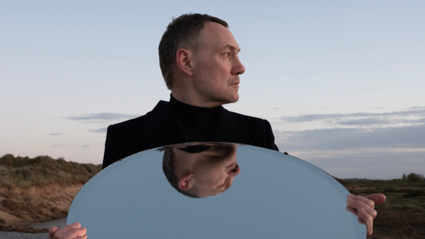image of David Gray holding a mirror underneath his face looking off to the distance