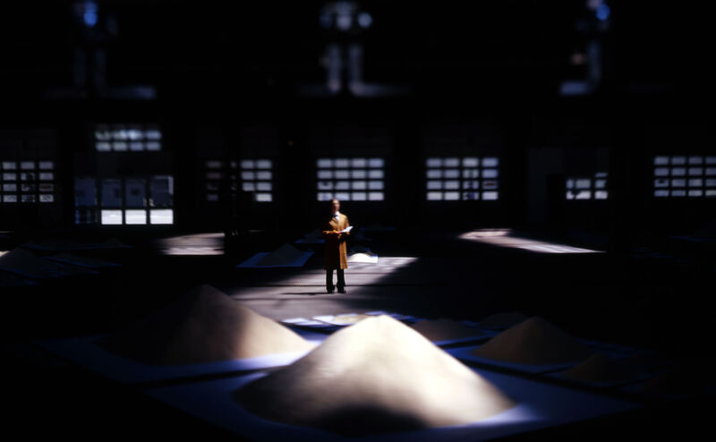 In a darkened room, a person is visible, standing amongst large piles of rice, lit by spotlights