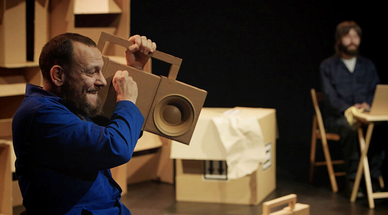 A man in a blue jumpsuit holds a cardboard boombox up to his ear and pretends to turn up the volume