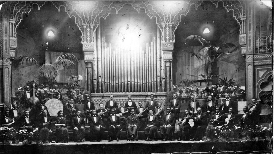 A large orchestra on the Dome stage with a big organ and plants pictured in the background