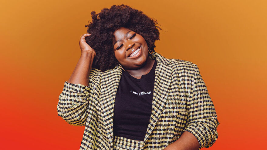 Stephanie Yeboah smiling, wearing a black t-shirt with white writing 'I Am Enough' and a yellow and black checked blazer. The background is a gradient from yellow at the top to red at the bottom