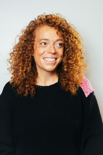 Michelle Wolf staring to the right smiling. She is wearing a black jumper