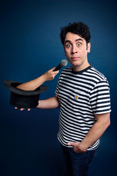 Pete Firman holding a top hat which has a hand and a microphone coming out of it. He has a surprised facial expression