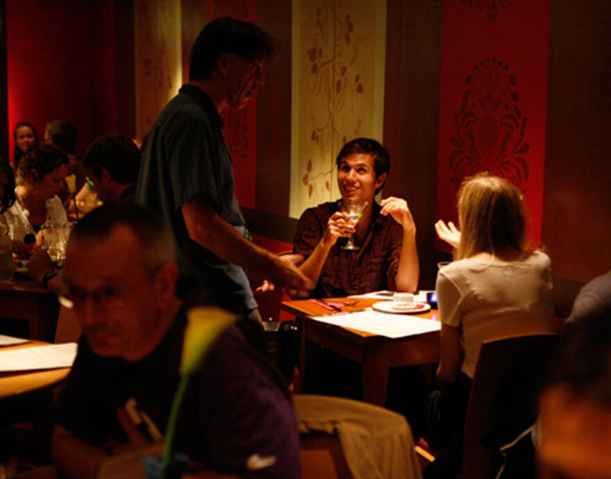 Restaurant interior, two people are sat at a table, light shining on them, as a waiter comes to take their order.