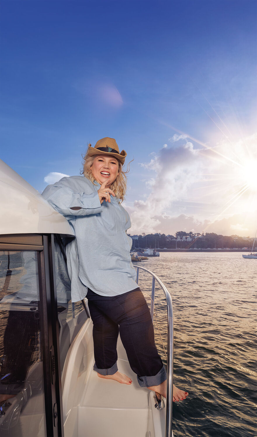 Urzila Carlson wearing a brown hat and a blue shirt standing on the side of yacht. The sun is shining and she has her hand to her chin