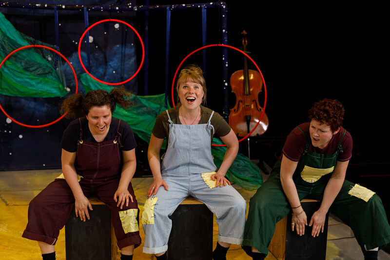 Three people sit on box drums, playing them and singing