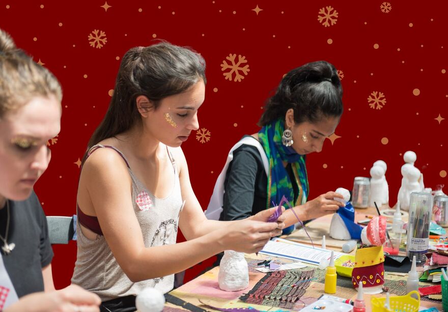 Three woman crafting at a table. They are in front of a red background with gold snowflakes