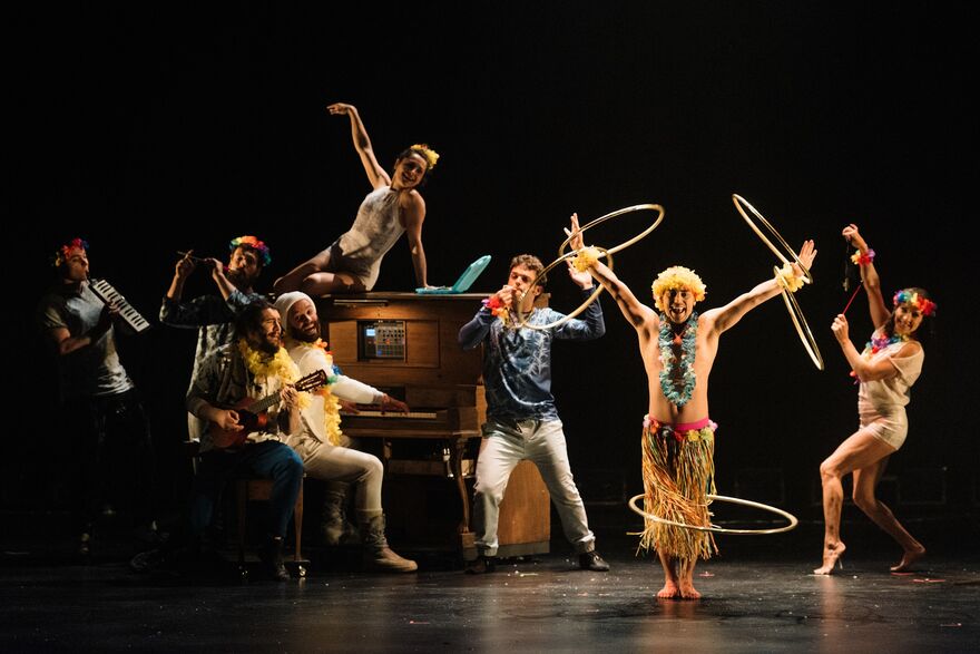 A group of performers posed around and on a piano whilst someone dressed as a hula girl is hula hooping in front of them all