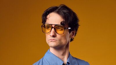 A young man with dark brown hair wearing a blue shirt and orange tinted sunglasses with an orange background