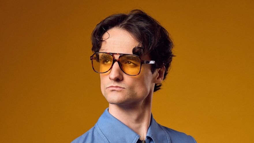 A young man with dark brown hair wearing a blue shirt and orange tinted sunglasses with an orange background