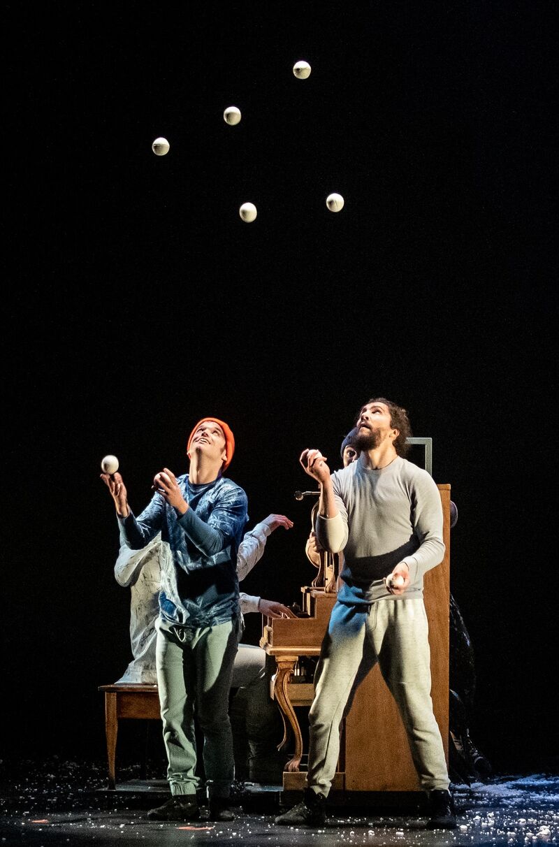 Two people on stage juggling white balls, stood in front of a brown piano