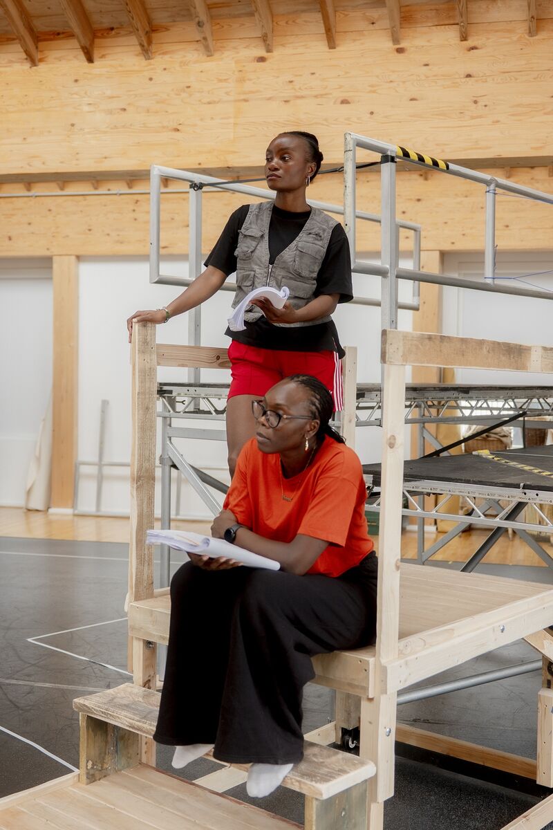 A black woman is sat on some wooden steps, she holds a script in her hands. Stood behind her on a wooden platform is another black woman, also holding a script. They are both dressed in black and red outfits.