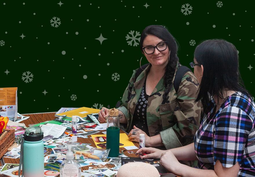 Two women looking at each other smiling. They are sat at a table full of crafts. The image is over a green background with snowflakes